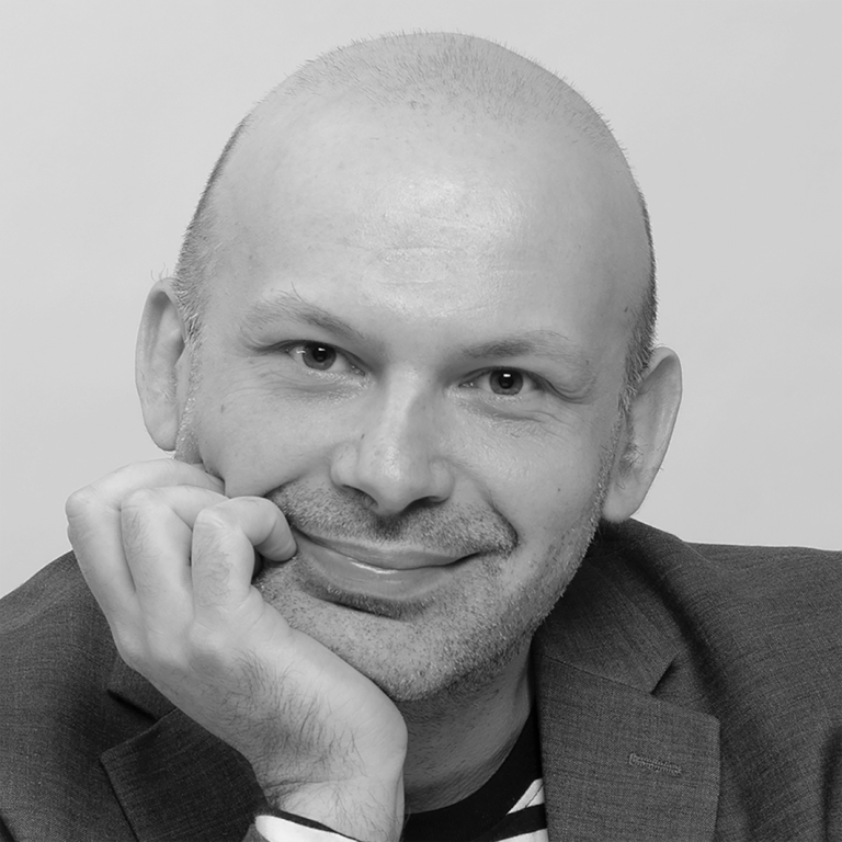 Czarno-białe zdjęcie - portret dr Grzegorza Dutki (niemający włosów mężczyzna w średnim wieku, głowa oparta na dłoni, szeroki uśmiech na twarzy)
