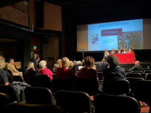 Sala konferencyjna. Na pierwszym planie plecy osób siedzących na widowni. W głębi - prezentacja wyświetlona na ekranie. Pod ekranem na scenie - stół pokryty obrusem przy którym siedzi 5 osób