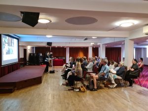 Sala konferencyjna. Widok z boku na scenę, ekran z prezentacją, prelegenta i widownię.