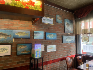 Wnętrze restauracji. Na ścianie z cegieł wisi 11 obrazów Artura Skowrona przedstawiających ryby morskie. W prawym dolnym roku stolik i dwa krzesła