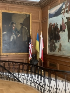 Wnętrze pomieszczenia w historycznym budynku. W rogu flagi: polska, amerykańska i ukraińska. Na ścianie po lewej stronie wisi bardzo duży obraz przedstawiający dwóch mężczyzn. Na ścianie po prawej stronie grupa osób ubranych w futra maszerujących w śniegu.