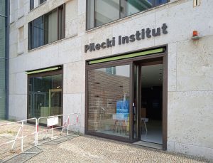 Fasada nowoczesnego budynku z otwartymi drzwiami. Nad drzwiami szyld "Pilecki Institut"