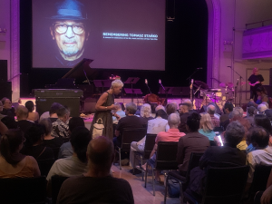 Wnętrze sali koncertowej. Nad sceną duży ekran z twarzą mężczyzny w okularach i kapeluszu i napisem "Remembering Tomasz Stańko". Poniżej fortepian i ustawione instrumenty muzyczne. Poniżej siedzenia wypełnione publicznością.